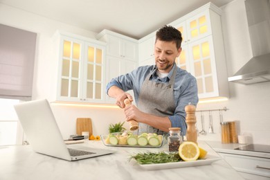 Photo of Man adding spices to dish while watching online cooking course via laptop in kitchen