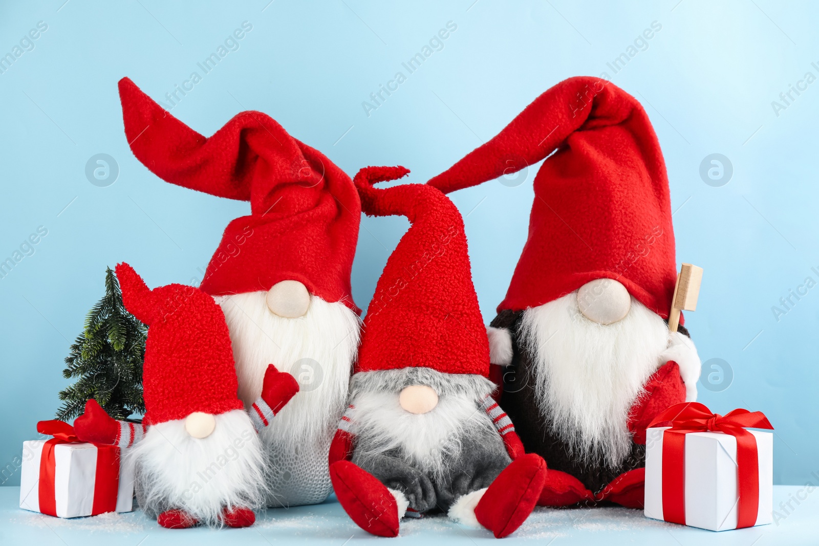 Photo of Many cute Christmas gnomes, gift boxes and small decorative fir tree on light blue background