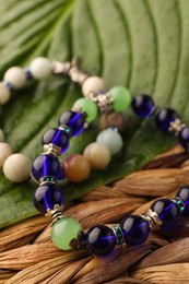 Beautiful bracelets with gemstones and green leaf on wicker surface, closeup