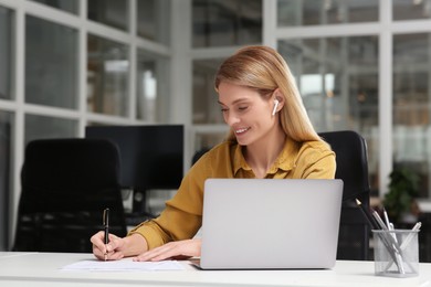 Photo of Woman writing notes while working on laptop at white desk in office