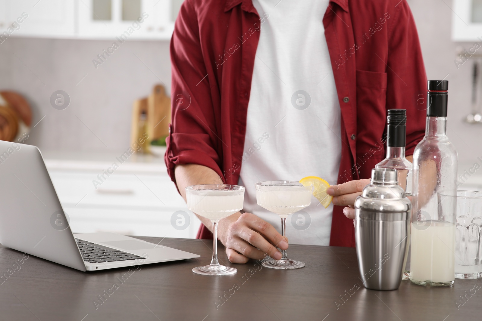 Photo of Man learning to make cocktail with online video on laptop at wooden table in kitchen, closeup. Time for hobby