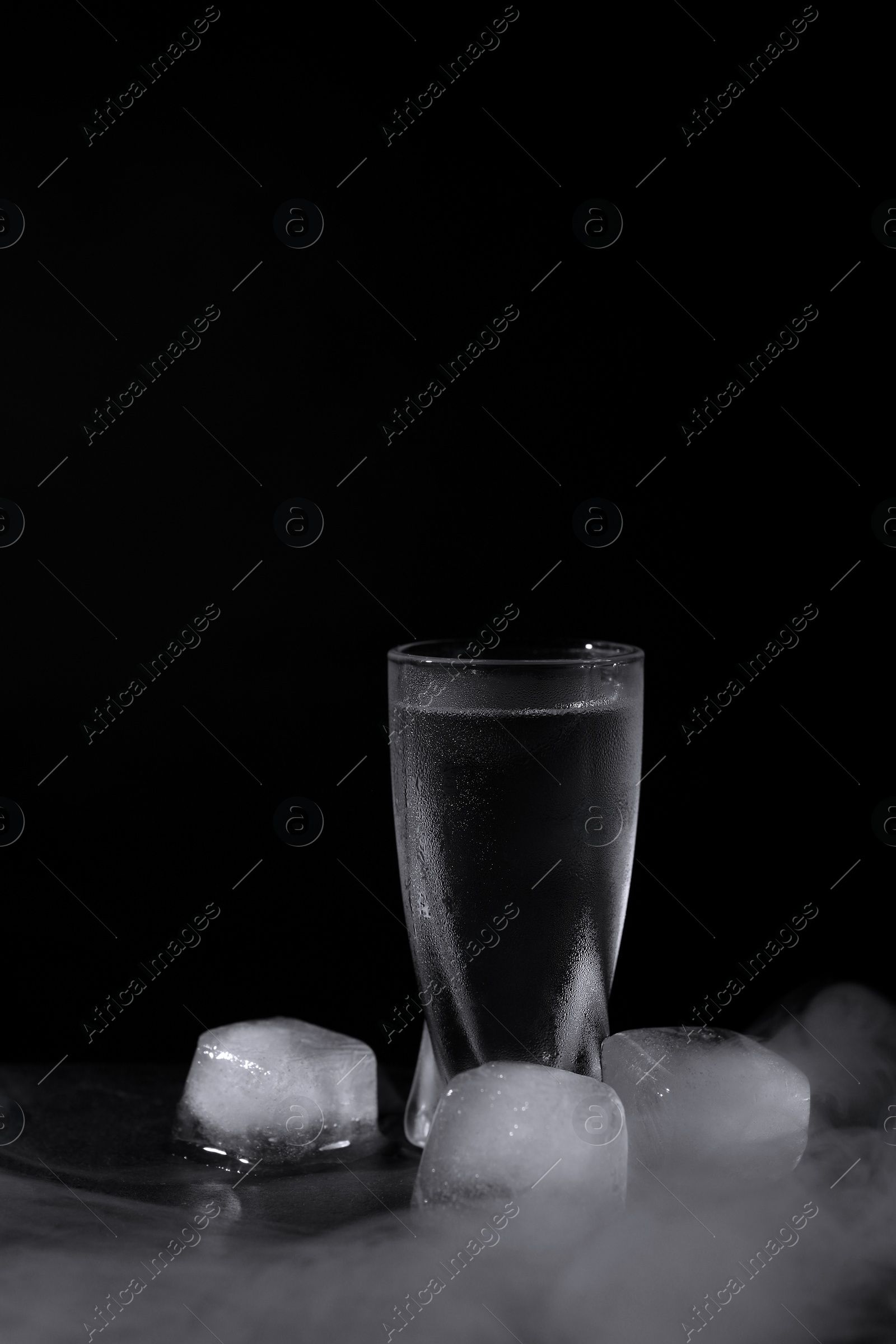 Photo of Vodka in shot glass with ice on table against black background