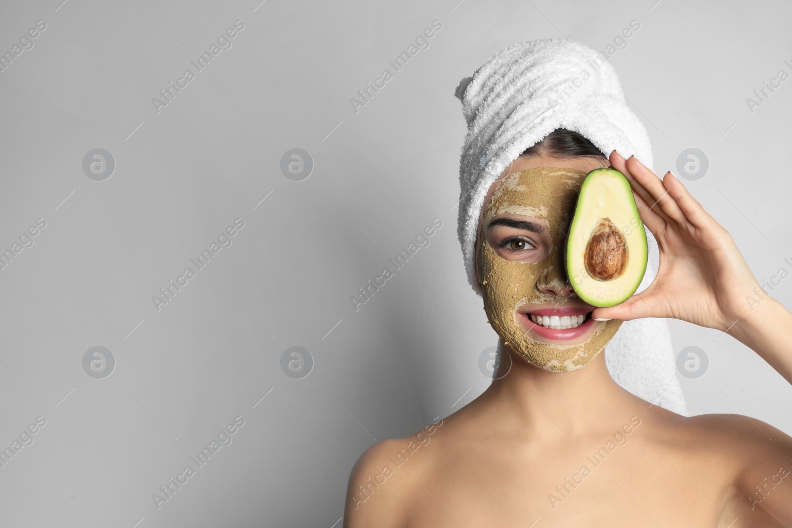 Photo of Young woman with clay mask on her face holding avocado against light background, space for text. Skin care