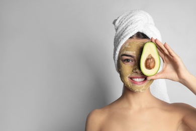 Photo of Young woman with clay mask on her face holding avocado against light background, space for text. Skin care