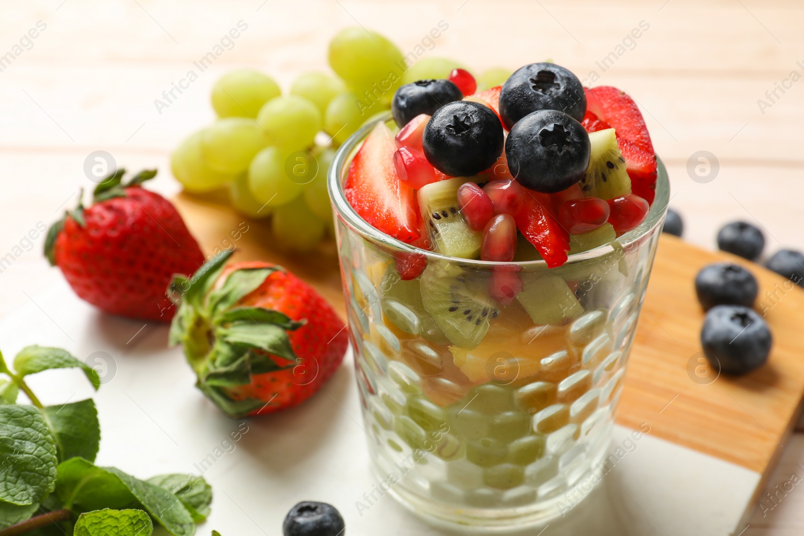 Photo of Healthy breakfast. Delicious fruit salad in glass and ingredients on light wooden table, closeup