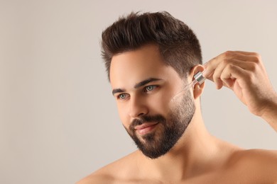 Handsome man applying cosmetic serum onto face on light grey background, space for text