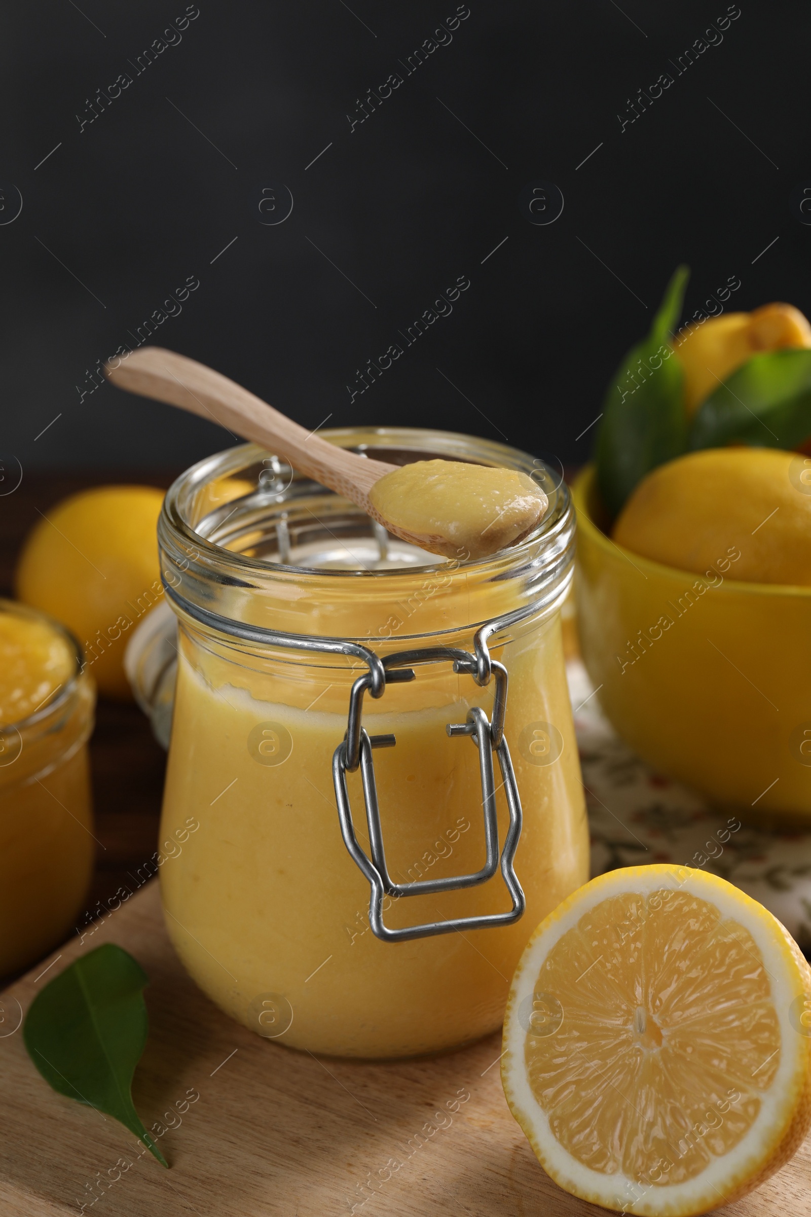 Photo of Delicious lemon curd in glass jar, spoon, fresh citrus fruits and green leaves on table