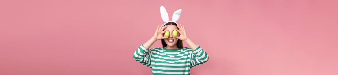 Image of Happy woman with bunny ears holding Easter eggs near eyes on pink background. Banner design