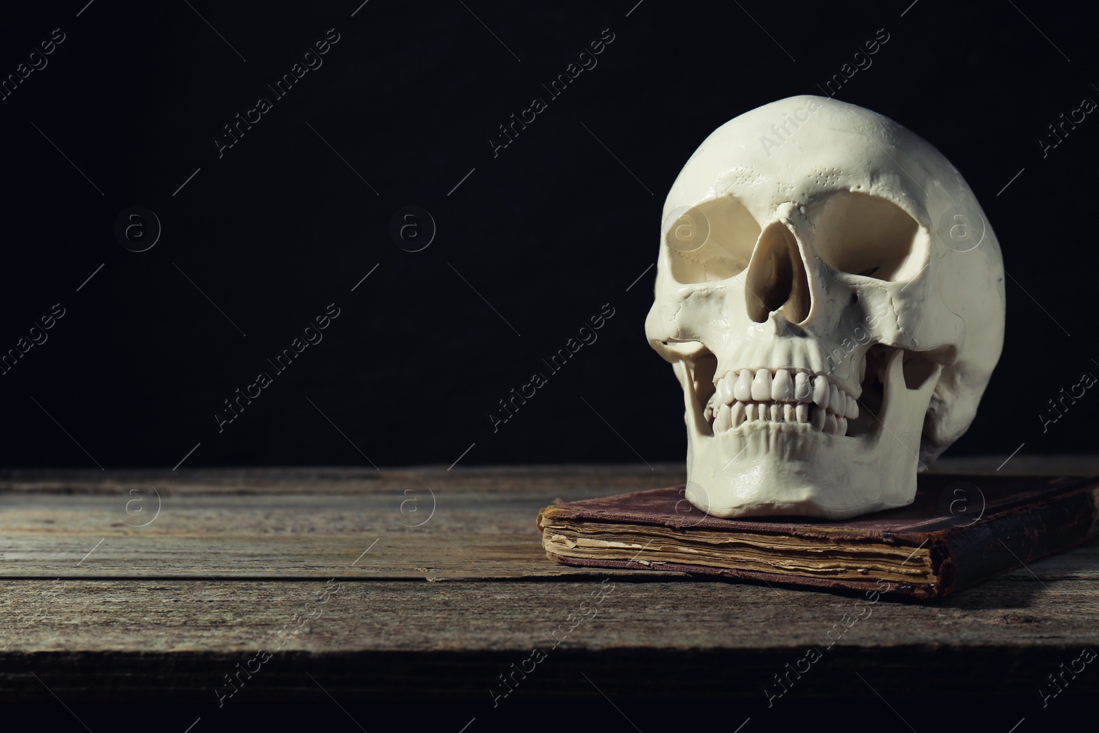 Photo of Human skull and old book on wooden table against black background, space for text