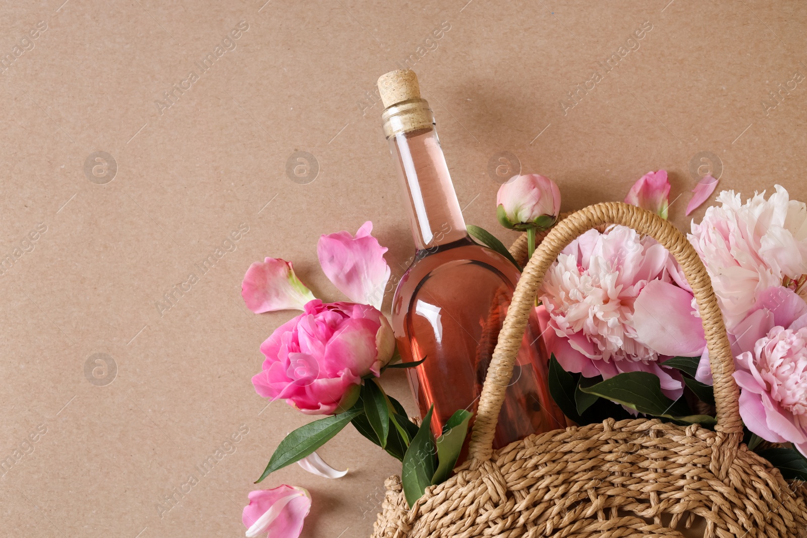 Photo of Wicker bag with bottle of rose wine and beautiful pink peonies on brown background, top view. Space for text