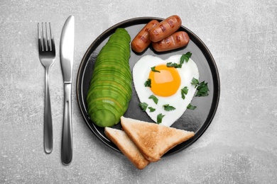 Romantic breakfast with fried heart shaped egg, avocado, sausages and toasts on light grey table, flat lay. Valentine's day celebration