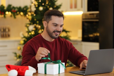 Photo of Celebrating Christmas online with exchanged by mail presents. Man opening gift box during video call on laptop at home