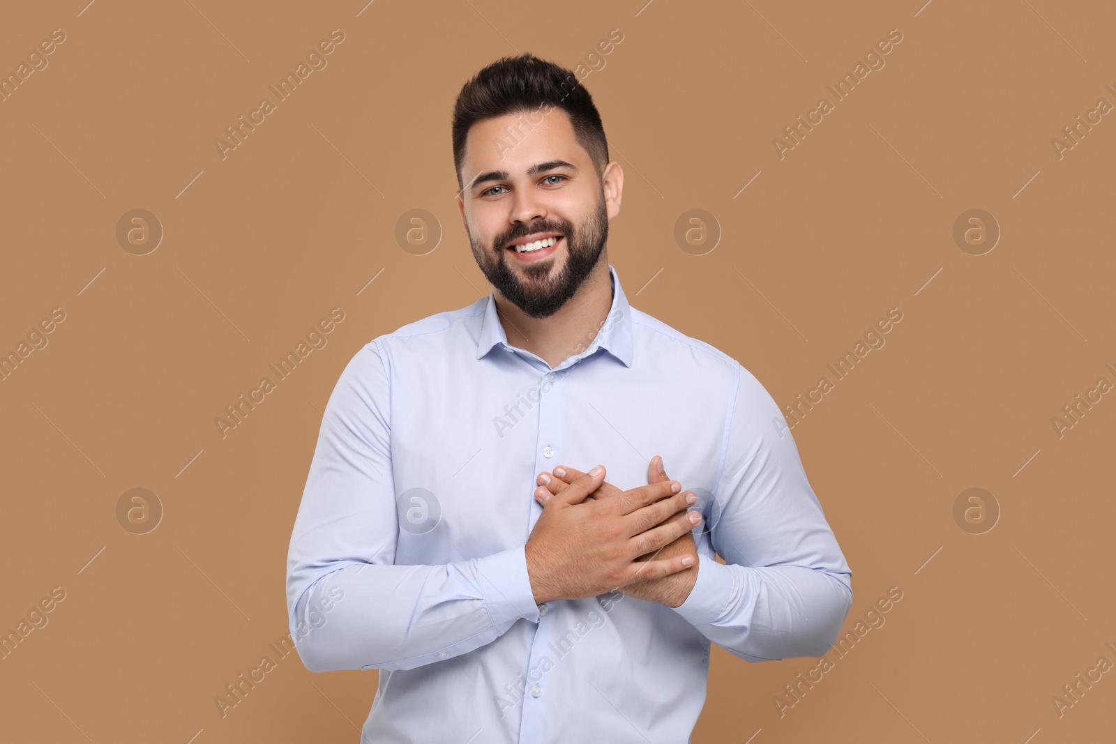 Photo of Thank you gesture. Handsome grateful man holding hands near heart on brown background