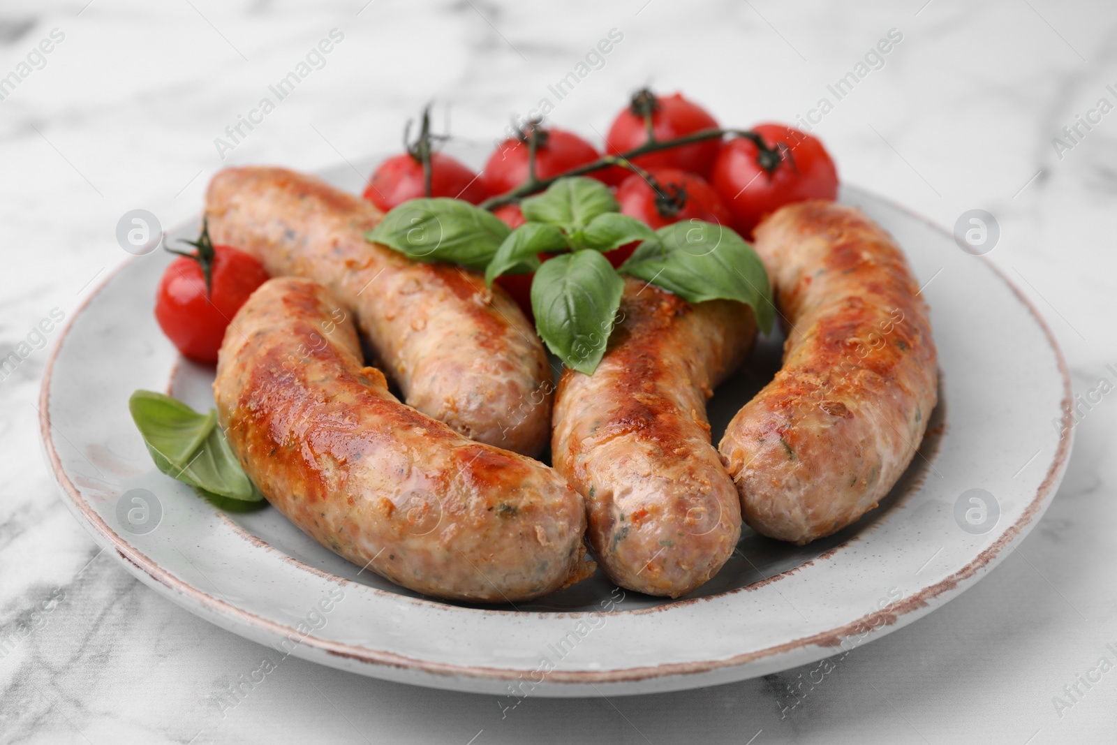 Photo of Plate with tasty homemade sausages, basil leaves and tomatoes on white marble table, closeup