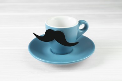 Photo of Cup with fake paper mustache and saucer on white wooden table