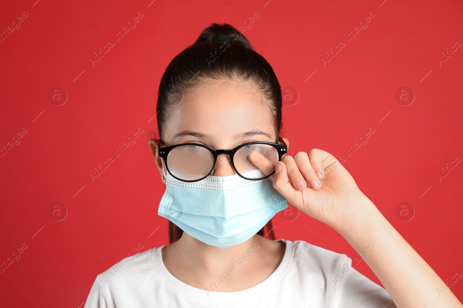 Photo of Little girl wiping foggy glasses caused by wearing medical face mask on red background. Protective measure during coronavirus pandemic