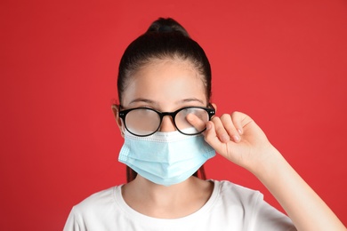 Little girl wiping foggy glasses caused by wearing medical face mask on red background. Protective measure during coronavirus pandemic