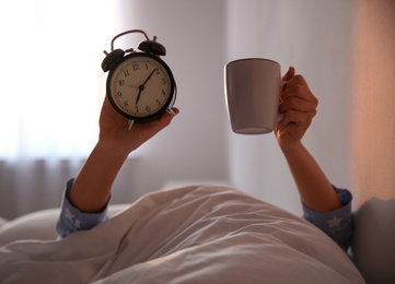 Woman with cup and alarm clock lying in bed, closeup. Morning time