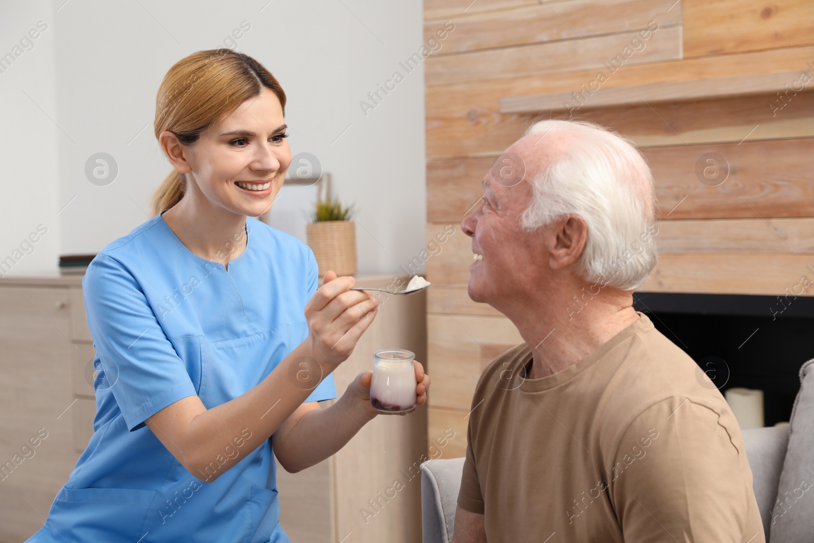 Photo of Nurse feeding elderly man with yogurt indoors. Assisting senior people