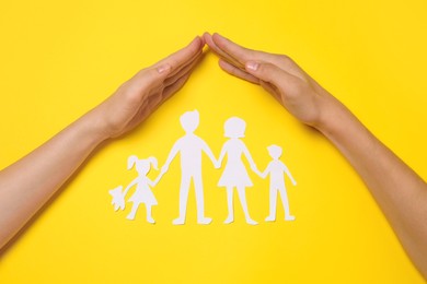 Photo of Woman and man covering paper family figures with hands on yellow background, top view