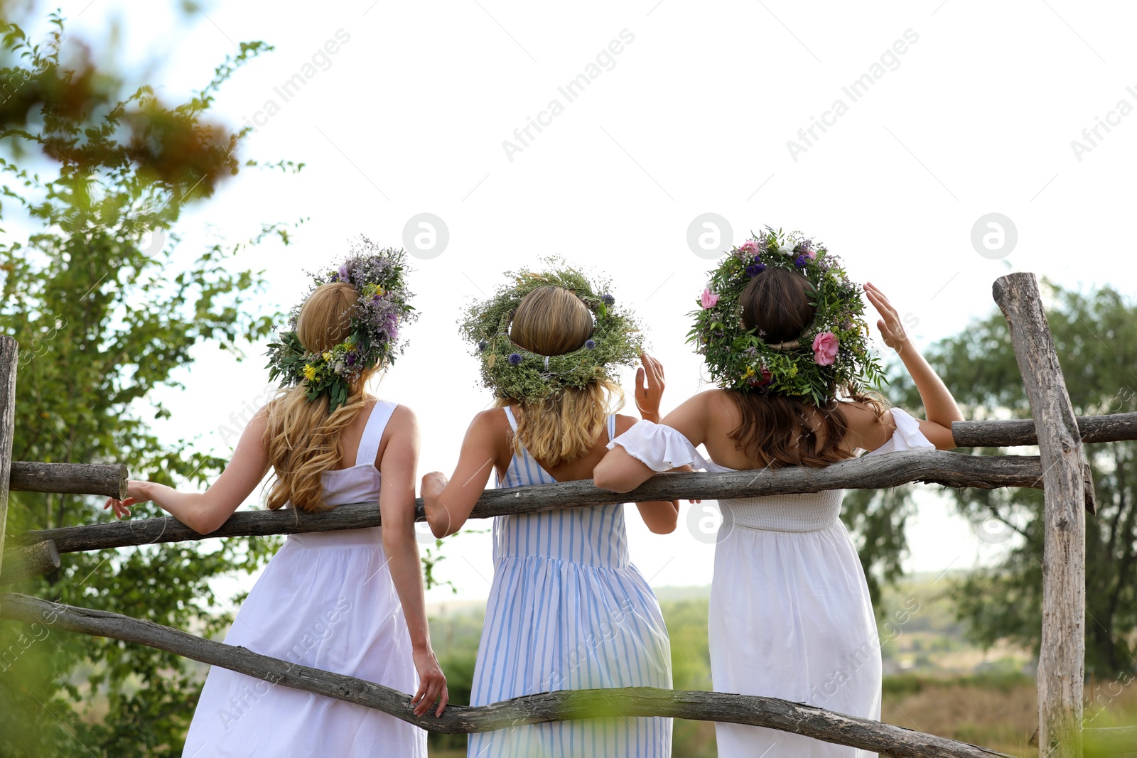 Photo of Young women wearing wreaths made of beautiful flowers near wooden fence, back view