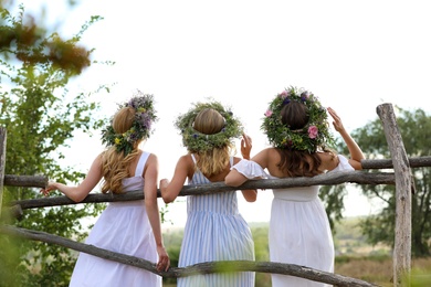 Young women wearing wreaths made of beautiful flowers near wooden fence, back view