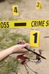 Crime scene with dead woman's body, bloody knife, markers and yellow tape outdoors, closeup