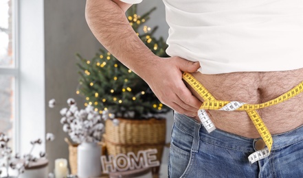 Overweight man measuring his waist in room decorated for Christmas after holidays, closeup