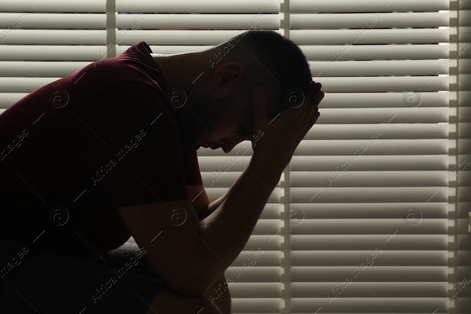 Photo of Sad man sitting near closed blinds indoors. Space for text