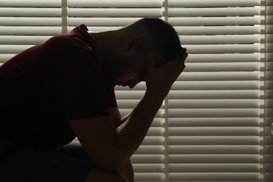 Photo of Sad man sitting near closed blinds indoors. Space for text