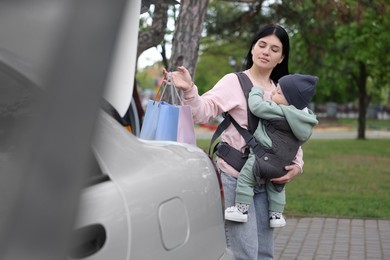 Mother holding her child in sling (baby carrier) while putting shopping bags into car trunk outdoors