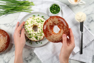 Photo of Woman making bagel with cream cheese and green onion at white marble table, top view