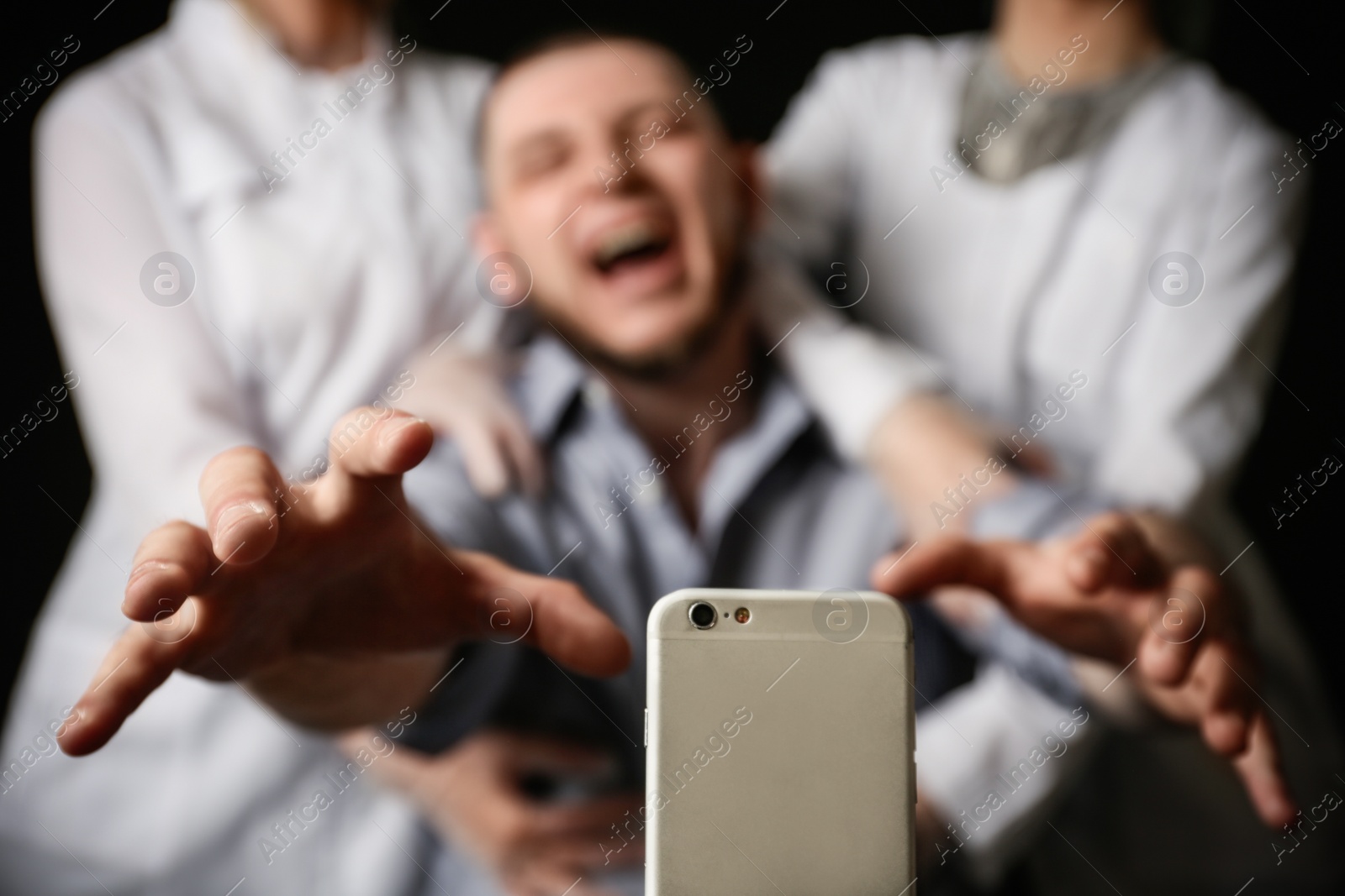 Photo of Emotional man reaching for smartphone while medical workers holding him back on black background, closeup. Addiction concept