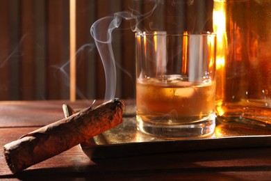Photo of Tray with smoldering cigar and whiskey on wooden table, closeup
