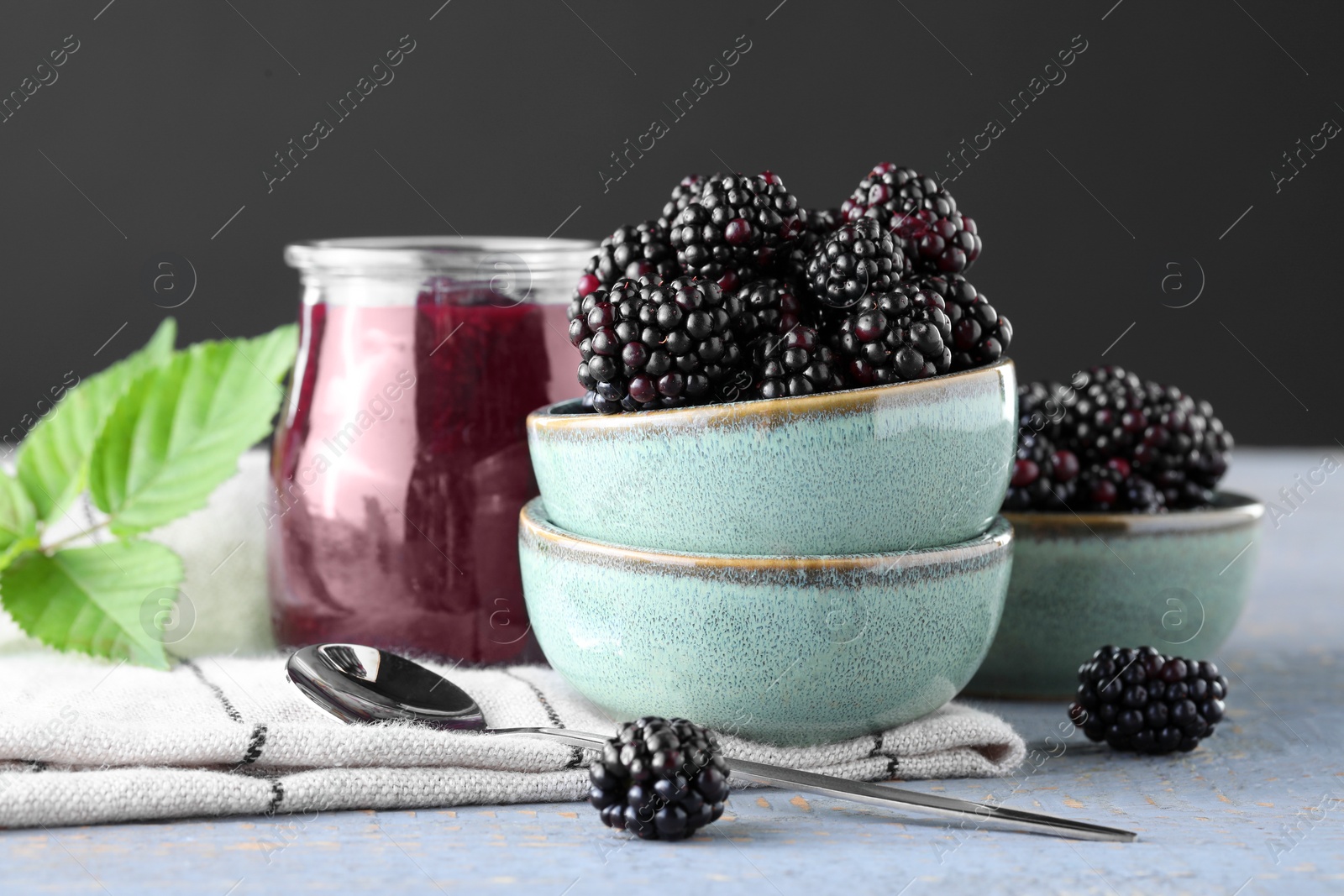 Photo of Fresh ripe blackberries, tasty jam and leaves on light grey wooden table