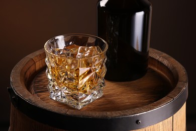 Whiskey with ice cubes in glass and bottle on wooden barrel, closeup