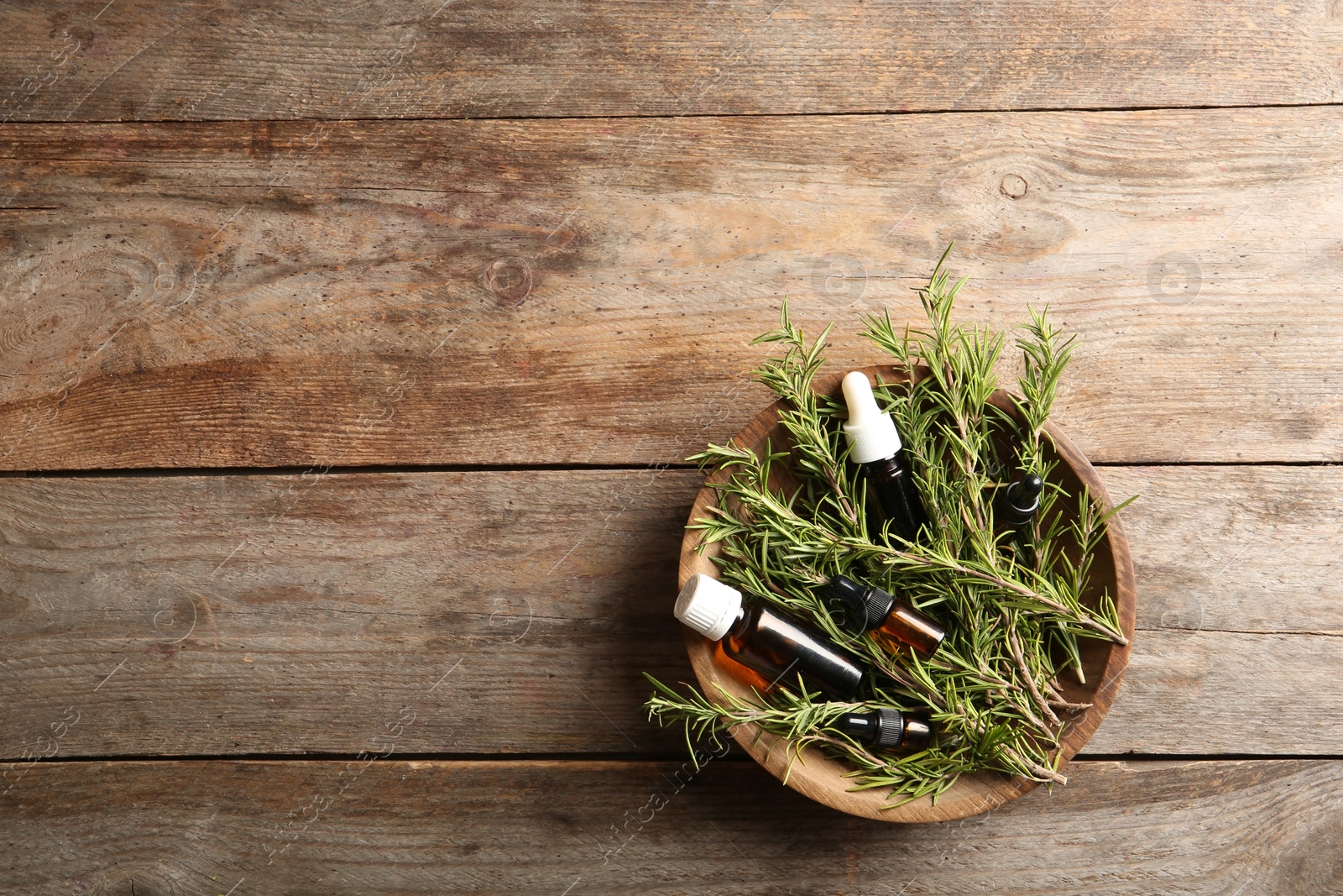 Photo of Bowl with bottles of rosemary oil and fresh twigs on wooden background, top view