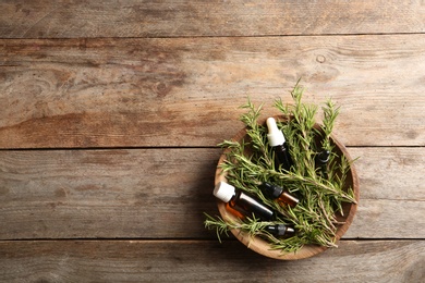 Photo of Bowl with bottles of rosemary oil and fresh twigs on wooden background, top view