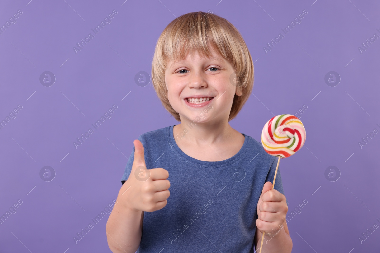 Photo of Happy little boy with colorful lollipop swirl showing thumbs up on violet background