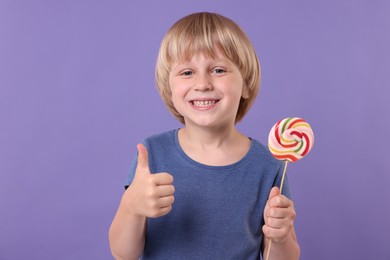 Happy little boy with colorful lollipop swirl showing thumbs up on violet background