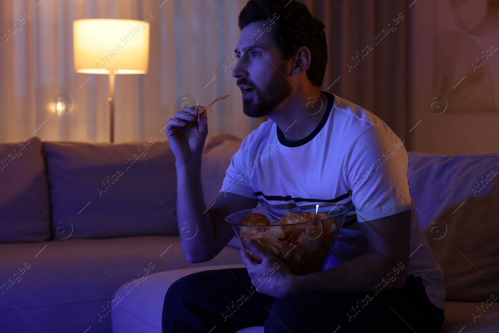 Photo of Man eating chips while watching TV on sofa at night. Bad habit