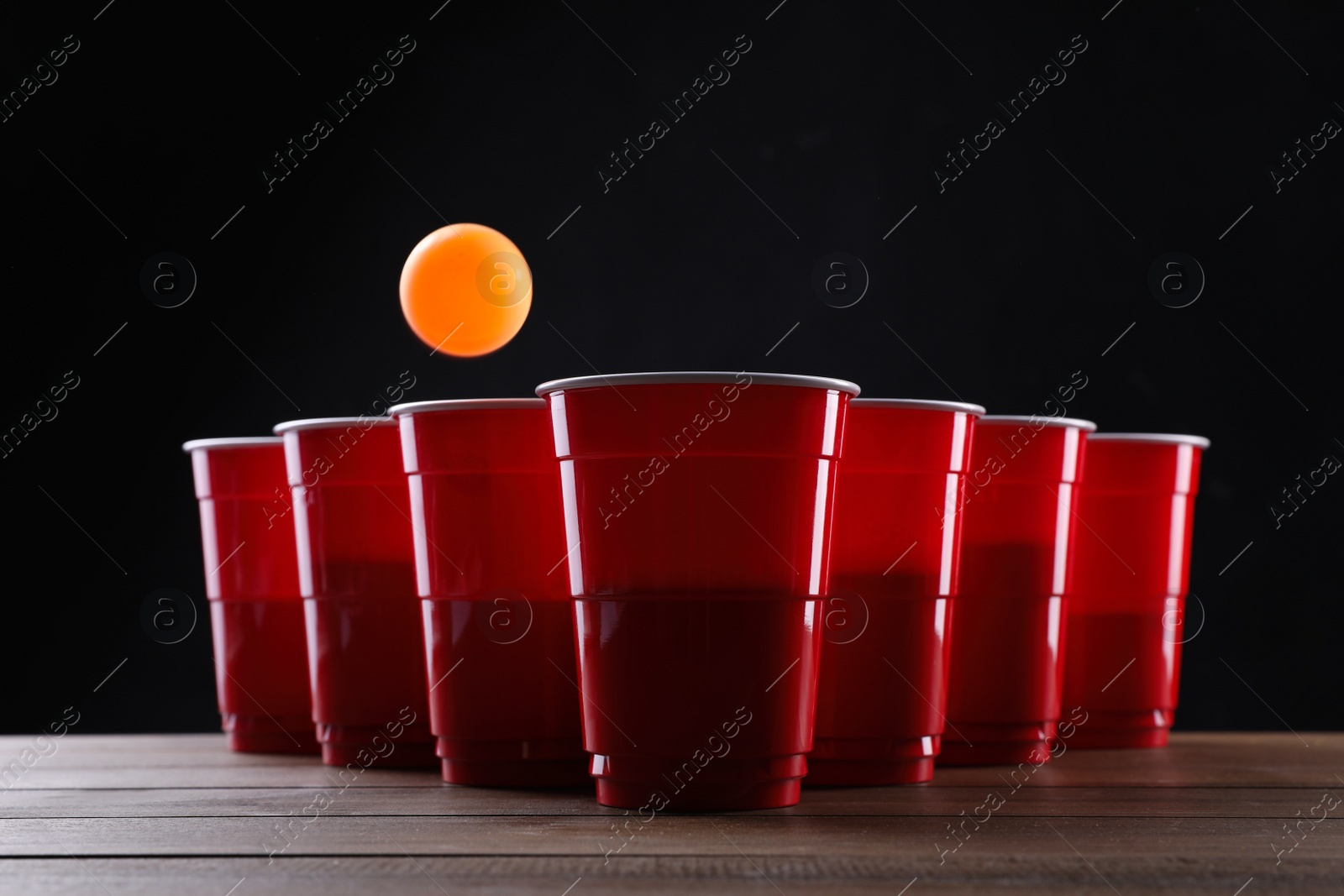 Photo of Plastic cups and ball for beer pong on wooden table against black background
