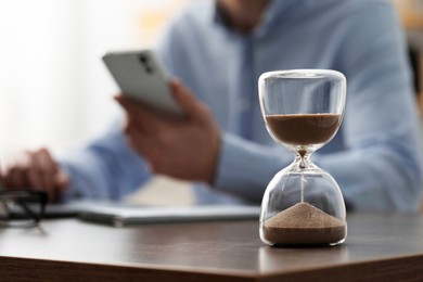 Photo of Hourglass with flowing sand on desk. Man using smartphone indoors, selective focus