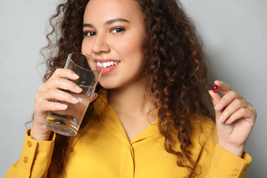 African-American woman with glass of water and vitamin capsule on light grey background