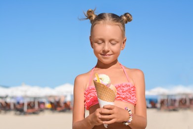 Photo of Adorable little girl in swimsuit with delicious ice cream at beach on sunny summer day