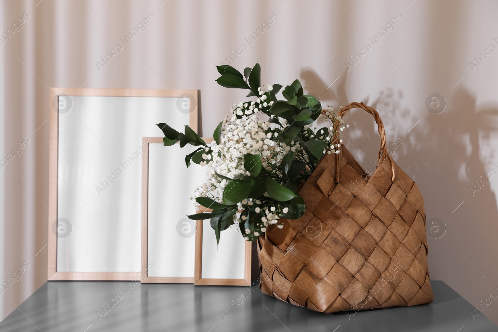 Photo of Stylish wicker basket with bouquet of flowers near empty frames on wooden table indoors