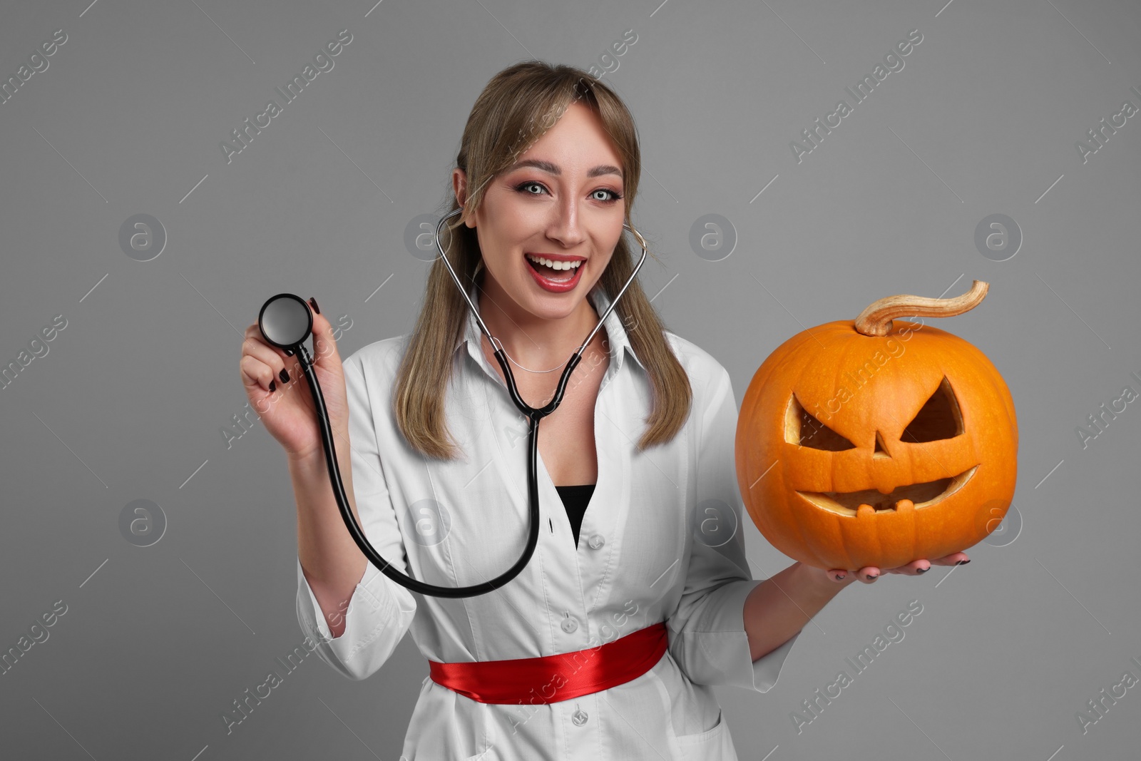 Photo of Woman in scary nurse costume with carved pumpkin and stethoscope on light grey background. Halloween celebration