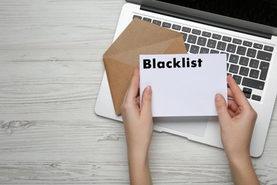 Woman holding letter with word Blacklist near laptop at white wooden table, top view. Space for text