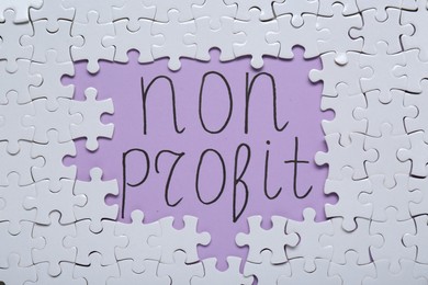 Photo of Phrase Non Profit surrounded by many white puzzles on purple background, flat lay