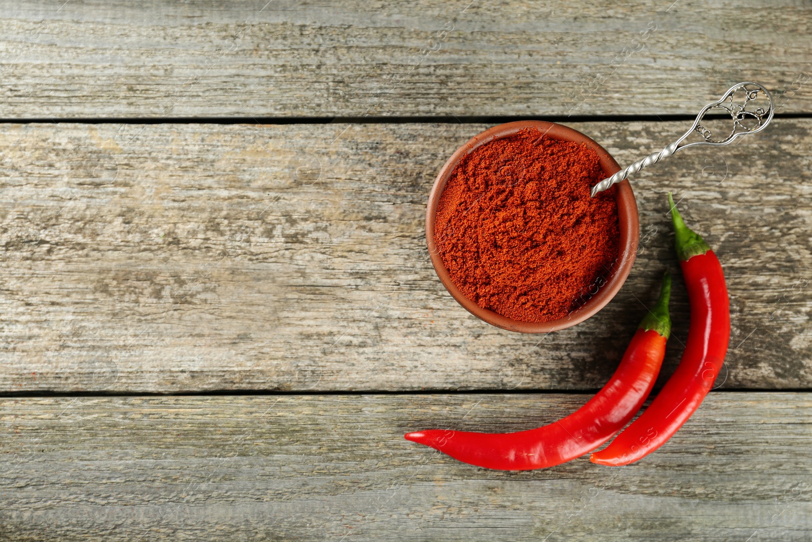 Photo of Bowl of ground red pepper with spoon and ingredient on wooden table, flat lay. Space for text